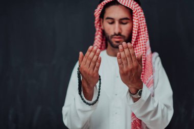 Arabian man in traditional clothes making traditional prayer to God, keeps hands in praying gesture in front of black chalkboard representing modern islam fashion and ramadan kareem concept. 