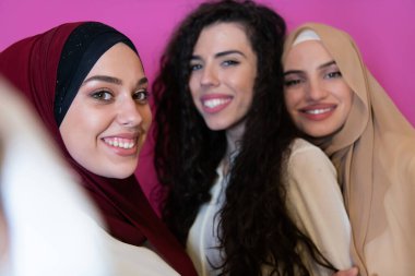 Group of beautiful muslim women two of them in fashionable dress with hijab using mobile phone while taking selfie picture isolated on pink background representing modern islam fashion technology.