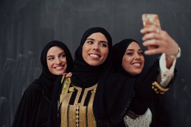 Group of young beautiful muslim women in fashionable dress with hijab using smartphone while taking selfie picture in front of black background. 