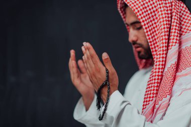 Arabian man in traditional clothes making traditional prayer to God, keeps hands in praying gesture in front of black chalkboard representing modern islam fashion and ramadan kareem concept. 