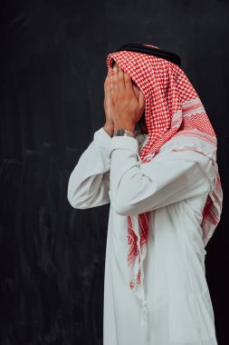 Arabian man in traditional clothes making traditional prayer to God, keeps hands in praying gesture in front of black chalkboard representing modern islam fashion and ramadan kareem concept. 