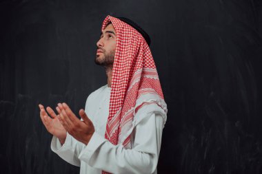 Arabian man in traditional clothes making traditional prayer to God, keeps hands in praying gesture in front of black chalkboard representing modern islam fashion and ramadan kareem concept. 