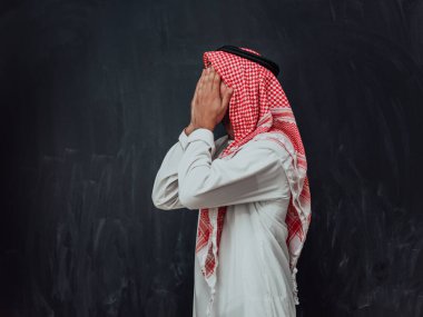 Arabian man in traditional clothes making traditional prayer to God, keeps hands in praying gesture in front of black chalkboard representing modern islam fashion and ramadan kareem concept. 