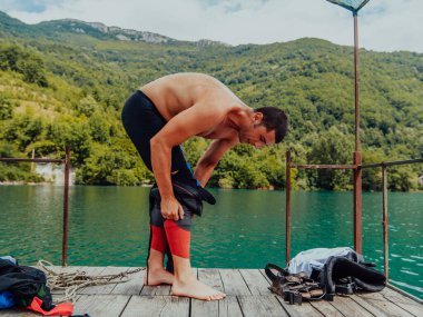 Athlete putting on a swimming suit and preparing for triathlon swimming and training in the river surrounded by natural greenery. 