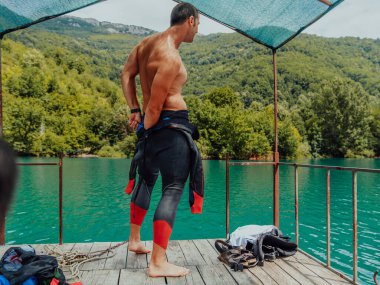 Athlete putting on a swimming suit and preparing for triathlon swimming and training in the river surrounded by natural greenery. 