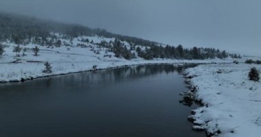 An aerial view of a frozen river flowing through snow-covered forests on a cloudy sunset sky background. Hi quality 4K footage.