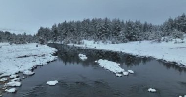 An aerial view of a frozen river flowing through snow-covered forests on a cloudy sunset sky background. Hi quality 4K footage.