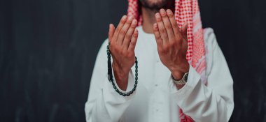 Arabian man in traditional clothes making traditional prayer to God, keeps hands in praying gesture in front of black chalkboard representing modern islam fashion and ramadan kareem concept. 