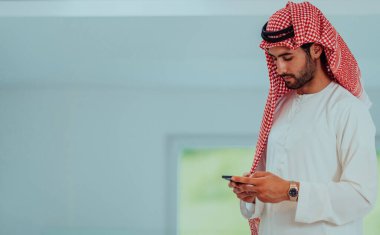 Young modern Muslim Arabian businessmen wearing traditional clothes while using smartphones at home