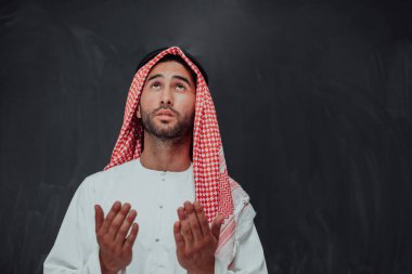 Arabian man in traditional clothes making traditional prayer to God, keeps hands in praying gesture in front of black chalkboard representing modern islam fashion and ramadan kareem concept. 