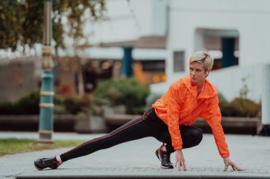Fit attractive woman in sportswear stretching one leg before jogging on the footpath outdoor in summer among greenery. Workout, sport, activity, fitness, vacation and training conept