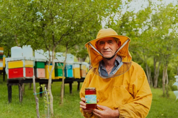 箱と蜂蜜の農場に囲まれた牧草地に立っている間 養蜂家は彼の手に蜂蜜の瓶を持っています — ストック写真