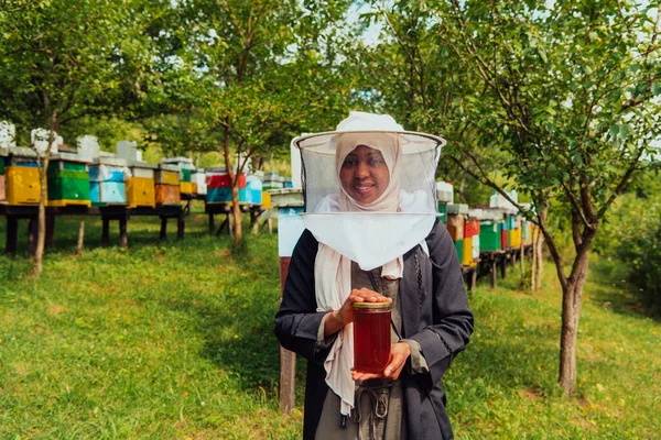 Portrait of a Muslim African American woman in the beekeeping department of a honey farm holding a jar of honey in her hand.