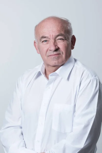 Portrait of an older man wearing a white shirt on a gray background. A healthy old man looks at a camera isolated over a gray wall. An older man smiles at the camera, a happy old man. High quality