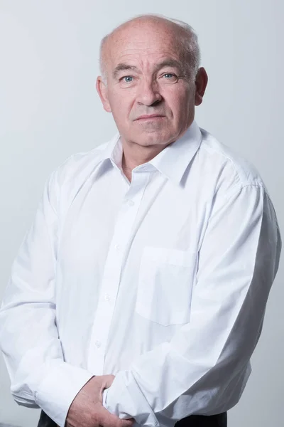 Portrait of an older man wearing a white shirt on a gray background. A healthy old man looks at a camera isolated over a gray wall. An older man smiles at the camera, a happy old man. High quality