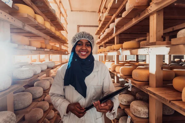 African American Muslim businesswoman checking product quality and entering data into a laptop at a local cheese manufacturing company.