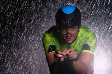 Gece boyunca bisiklet sürerken yağmura göğüs geren bir triatlet yaklaşan maratona hazırlanıyor. Ön plandaki bulanık yağmur damlaları ve arka plandaki karanlık, kasvetli atmosfer...