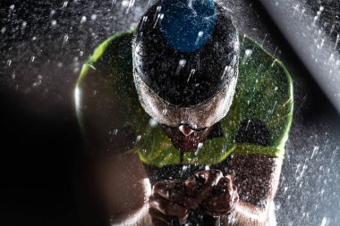 Gece boyunca bisiklet sürerken yağmura göğüs geren bir triatlet yaklaşan maratona hazırlanıyor. Ön plandaki bulanık yağmur damlaları ve arka plandaki karanlık, kasvetli atmosfer...