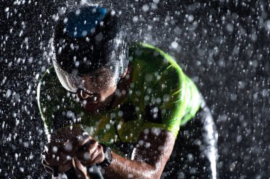 Gece boyunca bisiklet sürerken yağmura göğüs geren bir triatlet yaklaşan maratona hazırlanıyor. Ön plandaki bulanık yağmur damlaları ve arka plandaki karanlık, kasvetli atmosfer...