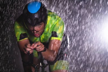 Gece boyunca bisiklet sürerken yağmura göğüs geren bir triatlet yaklaşan maratona hazırlanıyor. Ön plandaki bulanık yağmur damlaları ve arka plandaki karanlık, kasvetli atmosfer...