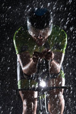 Gece boyunca bisiklet sürerken yağmura göğüs geren bir triatlet yaklaşan maratona hazırlanıyor. Ön plandaki bulanık yağmur damlaları ve arka plandaki karanlık, kasvetli atmosfer...