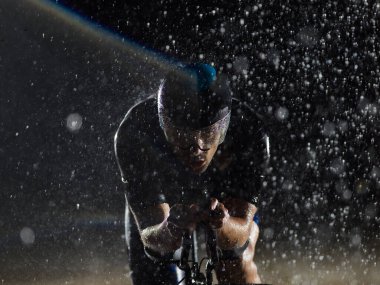 Gece boyunca bisiklet sürerken yağmura göğüs geren bir triatlet yaklaşan maratona hazırlanıyor. Ön plandaki bulanık yağmur damlaları ve arka plandaki karanlık, kasvetli atmosfer...