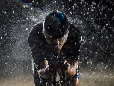 Gece boyunca bisiklet sürerken yağmura göğüs geren bir triatlet yaklaşan maratona hazırlanıyor. Ön plandaki bulanık yağmur damlaları ve arka plandaki karanlık, kasvetli atmosfer...