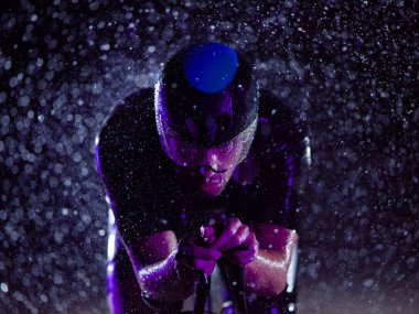 Gece boyunca bisiklet sürerken yağmura göğüs geren bir triatlet yaklaşan maratona hazırlanıyor. Ön plandaki bulanık yağmur damlaları ve arka plandaki karanlık, kasvetli atmosfer...