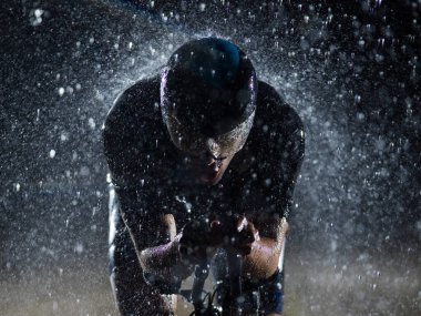 Gece boyunca bisiklet sürerken yağmura göğüs geren bir triatlet yaklaşan maratona hazırlanıyor. Ön plandaki bulanık yağmur damlaları ve arka plandaki karanlık, kasvetli atmosfer...