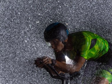 Gece boyunca bisiklet sürerken yağmura göğüs geren bir triatlet yaklaşan maratona hazırlanıyor. Ön plandaki bulanık yağmur damlaları ve arka plandaki karanlık, kasvetli atmosfer...