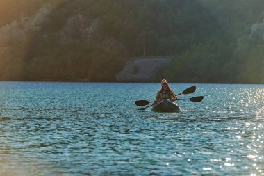 Genç bir çift, günbatımında orman yeşiliyle çevrili güzel bir nehrin ortasında kayak yolculuğunun tadını çıkarıyor..