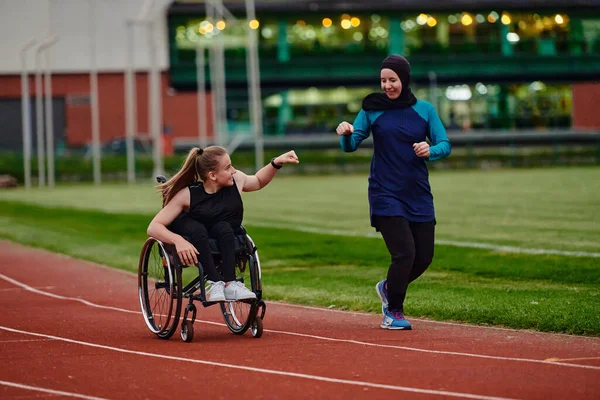 ブルカを身に着けているイスラム教徒の女性は マラソンコースで一緒に訓練する際に車椅子で障害のある友人をサポートしています — ストック写真