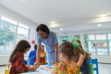 Bir kreşteki resim dersinde yaratıcı çocuklar ya da ilkokul sınıfında bayan öğretmenle resim çizen çocuklar.