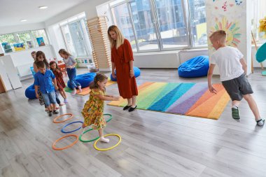 Küçük anaokulu öğrencileri ve bayan öğretmenleri sınıfın içinde egzersiz yapıyorlar. Hula hoop çemberinin üzerinden zıplarken yerde
