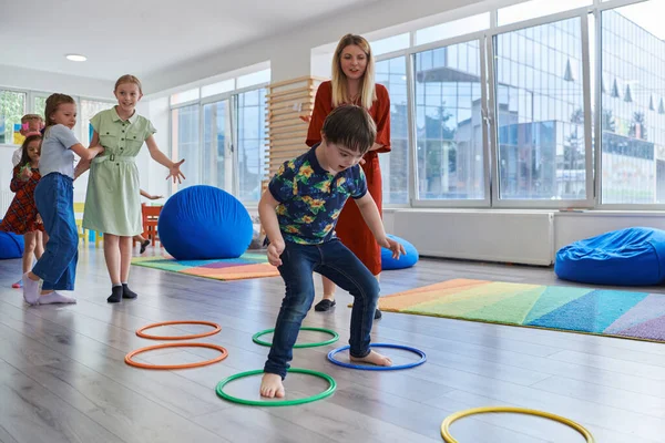 Small nursery school children with female teacher on floor indoors in classroom, doing exercise. Jumping over hula hoop circles track on the floor