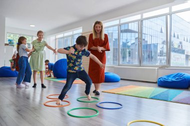 Küçük anaokulu öğrencileri ve bayan öğretmenleri sınıfın içinde egzersiz yapıyorlar. Hula hoop çemberinin üzerinden zıplarken yerde
