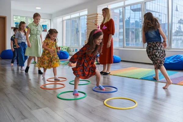 stock image Small nursery school children with female teacher on floor indoors in classroom, doing exercise. Jumping over hula hoop circles track on the floor