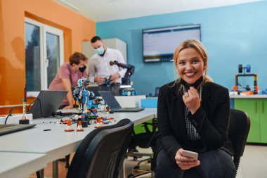 Bir kadın laboratuvarda oturup problemleri çözüyor ve robotların doğrulamasını inceliyor. Arka planda, meslektaşlar çevrimiçi bir toplantıda konuşuyor.. 