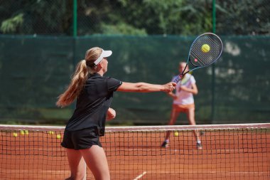 Genç kızlar güneşli bir günde canlı bir tenis maçında modern bir tenis kortunda yeteneklerini ve heveslerini gösteriyorlar.