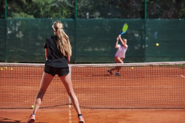 Genç kızlar güneşli bir günde canlı bir tenis maçında modern bir tenis kortunda yeteneklerini ve heveslerini gösteriyorlar.