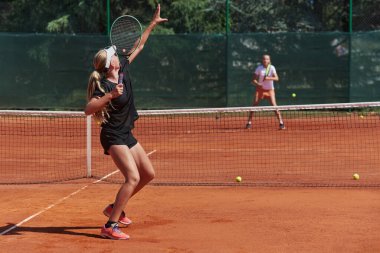 Genç kızlar güneşli bir günde canlı bir tenis maçında modern bir tenis kortunda yeteneklerini ve heveslerini gösteriyorlar.