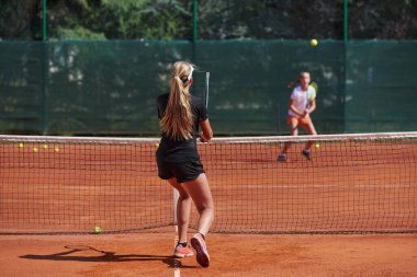 Genç kızlar güneşli bir günde canlı bir tenis maçında modern bir tenis kortunda yeteneklerini ve heveslerini gösteriyorlar.