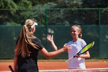 Güneşli bir günde gülümseyen iki bayan tenisçi, rekabetçi bir maçın ardından sportmenlik ve dostluk saçarak el sıkışıyor.