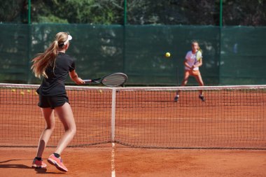 Genç kızlar güneşli bir günde canlı bir tenis maçında modern bir tenis kortunda yeteneklerini ve heveslerini gösteriyorlar.