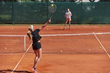 Genç kızlar güneşli bir günde canlı bir tenis maçında modern bir tenis kortunda yeteneklerini ve heveslerini gösteriyorlar.