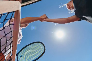 Güneşli bir günde gülümseyen iki bayan tenisçi, rekabetçi bir maçın ardından sportmenlik ve dostluk saçarak el sıkışıyor.