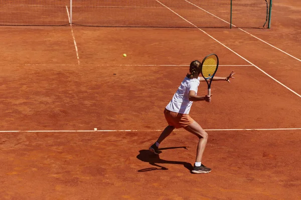 A young girl showing professional tennis skills in a competitive match on a sunny day, surrounded by the modern aesthetics of a tennis court
