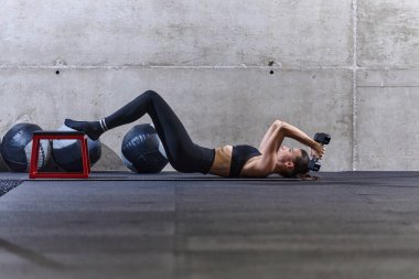 Formda bir kadın spor salonunda uzanıyor, dambıllarıyla kol egzersizleri yapıyor ve kendini adamışlığını ve gücünü gösteriyor.