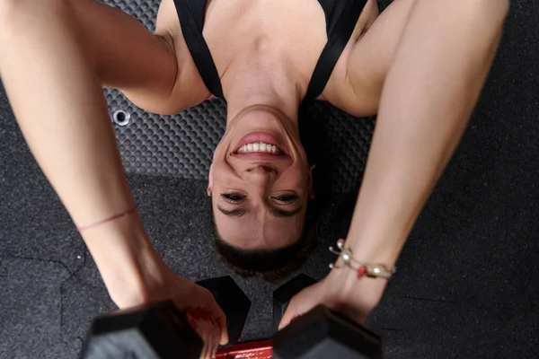一个健康的女人躺在体育馆的地板上 用哑铃进行手臂运动 展示她的奉献精神和力量 — 图库照片
