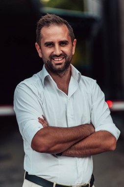 Beyaz gömlekli başarılı bir iş adamı, kollarını kavuşturmuş, açık havada poz veriyor, yüzünde kendinden emin bir ifade var.